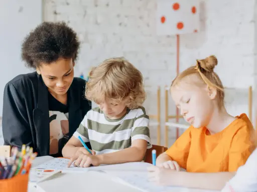 Kinder konzentriert bei Lernritualen in der Grundschule. Ein Mädchen mit schwarzer Afro-Frisur schaut zu, während ein Junge mit blonden Locken und ein Mädchen mit roten Haaren schreiben.