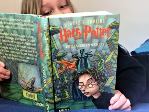 Ein Kind liest Harry Potter und die Kammer des Schreckens, eine Harry Potter Kinderversion. Der Zauberhut und ein Phönix sind auf dem Cover zu sehen.