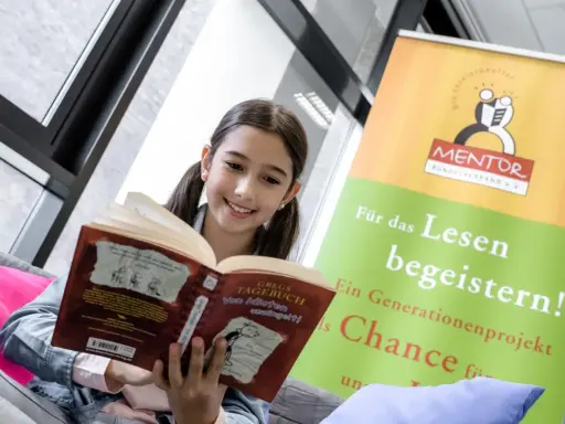 Ein lächelndes Mädchen liest ein Buch. Ein Banner im Hintergrund wirbt für "MENTOR" und "digitales lernen" durch Lesen.