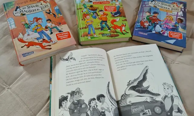 Drei Bücher der Reihe "Die Schule der magischen Tiere" von Margit Auer liegen nebeneinander. Aufgeschlagen ist ein Band mit einem Krokodil und einem Jungen.