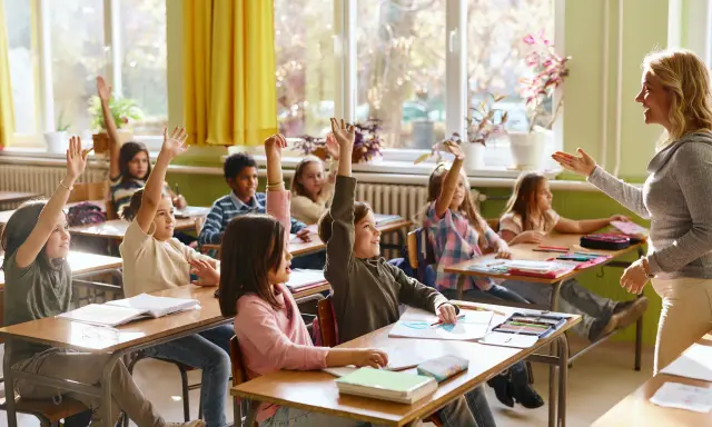 Grundschüler melden sich eifrig, ihre Sitzordnung zur Wand hin ausgerichtet, während die Lehrerin lächelt.