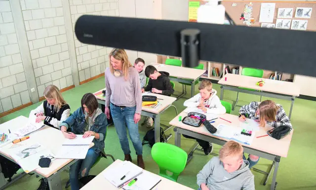 Lehrerin betreut Schüler im Klassenzimmer, die an ihren Tischen arbeiten. Ein modernes Setup für **hybrid unterricht**.