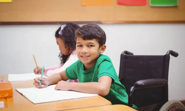 Ein Junge im Rollstuhl lächelt, während er im Unterricht schreibt. Dies ist ein Beispiel für **Inklusion in der Schule**.