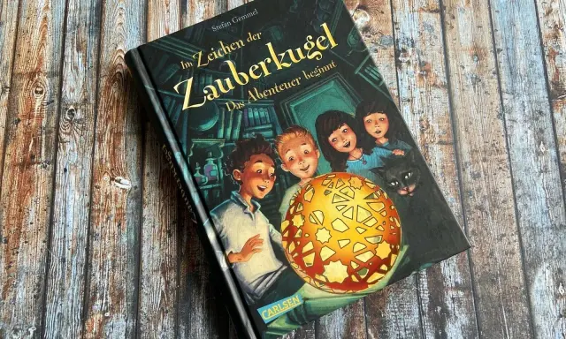 Ein Buchcover mit dem Titel "Im Zeichen der Zauberkugel". Kinder und eine Katze blicken neugierig auf eine leuchtende Kugel.