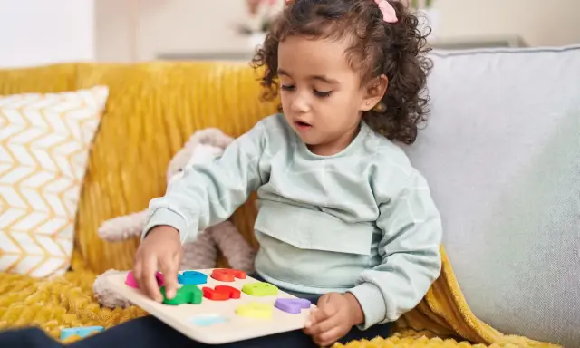 Kleines Mädchen mit Locken spielt konzentriert mit einem bunten Steckpuzzle. Das Spielzeug hilft, die **Konzentration zu steigern**.