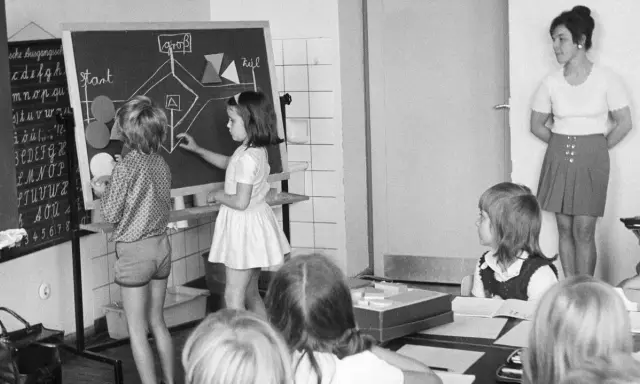 Unterricht in einer Schule um 1900. Kinder lernen an einer Tafel, während die Lehrerin zusieht.