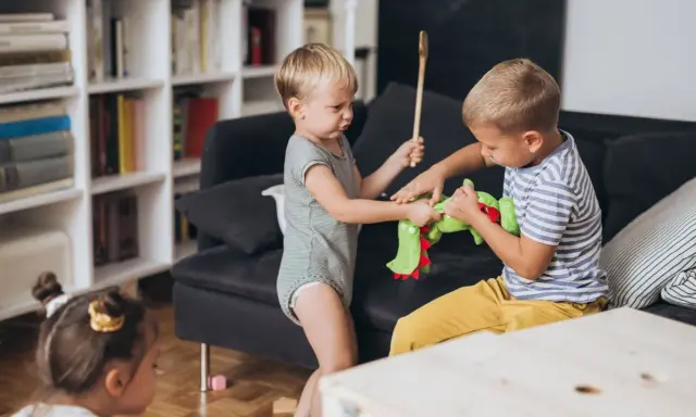 Zwei kleine Jungen streiten sich um ein grünes Stofftier. Einer hält einen Kochlöffel, der andere zieht am Spielzeug. Ein kleines Kind schaut zu.