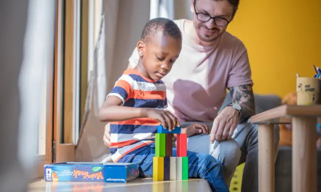 Ein Mann spielt mit einem Kind im Kindergarten. Der Junge baut einen Turm aus bunten Holzklötzen. Der Mann lächelt ihn an. Kein Trennungsschmerz im Kindergarten, nur Spiel und Spaß.