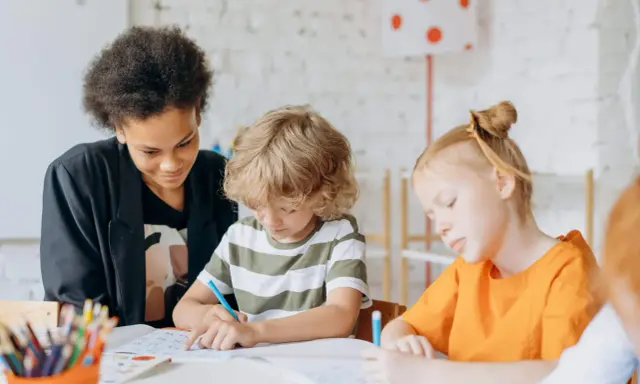 Kinder konzentriert bei Lernritualen in der Grundschule. Ein Mädchen mit schwarzer Afro-Frisur schaut zu, während ein Junge mit blonden Locken und ein Mädchen mit roten Haaren schreiben.