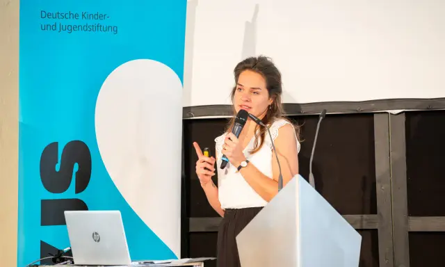 Eine Rednerin spricht über den Umgang mit Misserfolg. Im Hintergrund ein Banner der Deutschen Kinder- und Jugendstiftung.