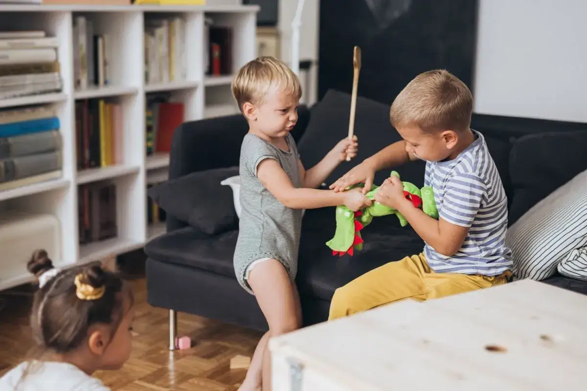 Zwei kleine Jungen streiten sich um ein gr&uuml;nes Stofftier. Einer h&auml;lt einen Kochl&ouml;ffel, der andere zieht am Spielzeug. Ein kleines Kind schaut zu.