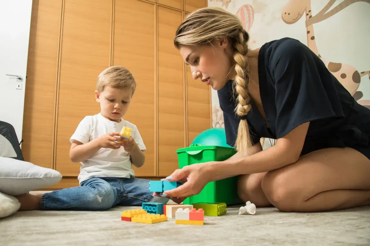 Mutter und Sohn bauen mit bunten Kl&ouml;tzen. Ein kleiner Junge h&auml;lt einen gelben Klotz, w&auml;hrend die Mutter ihm einen blauen gibt. Der Trennungsschmerz Kindergarten ist hier nicht zu sp&uuml;ren, nur Spiel und Spa&szlig;.