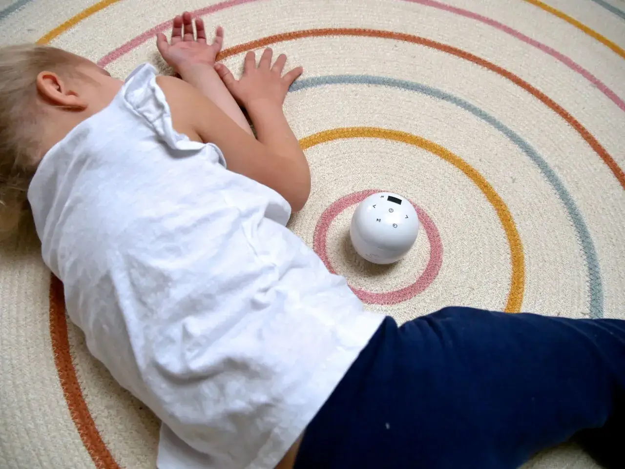 Kleines Kind liegt auf einem Teppich mit bunten Kreisen. Ein wei&szlig;er Ball mit Kn&ouml;pfen liegt daneben. Hilft reizoffene Kinder zur Ruhe zu kommen.