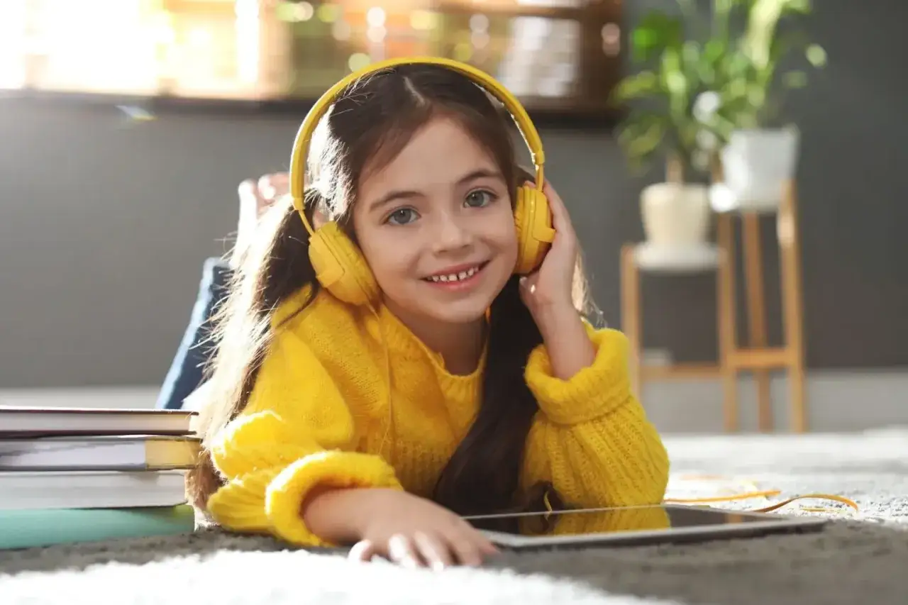 Ein l&auml;chelndes M&auml;dchen mit gelben Kopfh&ouml;rern h&ouml;rt aufmerksam zu. Neben ihr liegen B&uuml;cher und ein Tablet &ndash; perfekt f&uuml;r podcast englisch lernen kinder.