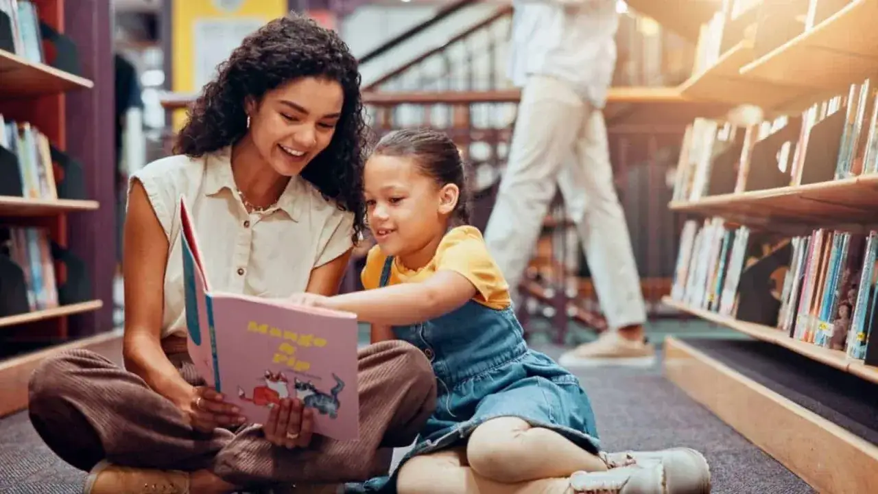 Eine Frau und ein Kind sitzen auf dem Boden einer Bibliothek und lesen ein Buch. Sie sind umgeben von B&uuml;cherregalen, was auf eine Atmosph&auml;re von Bewegung und Lernen hindeutet.