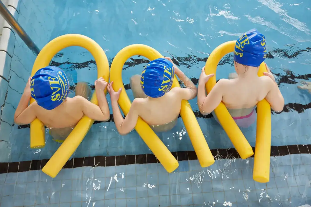 Drei Kinder mit Schwimmkappen &uuml;ben im Schwimmbad mit gelben Schwimmnudeln. Ein fr&ouml;hlicher Tag f&uuml;r das Schwimmen in der Grundschule.