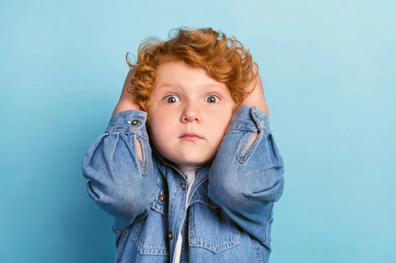 Ein Junge mit roten Haaren h&auml;lt sich die Ohren zu, seine Augen sind weit aufgerissen. Er wirkt &uuml;berfordert, ein Zeichen f&uuml;r motorische Unruhe bei Kindern.