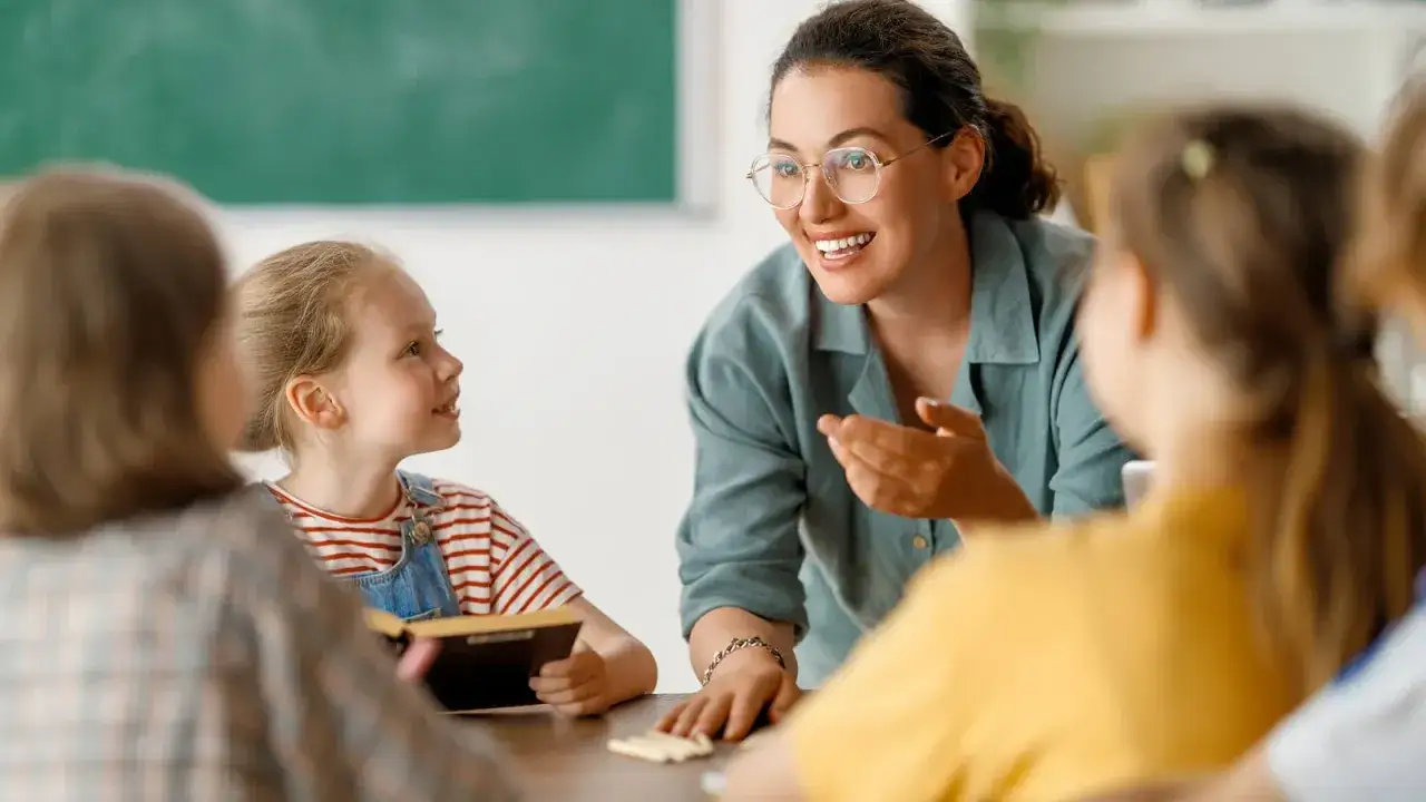 Lehrerin erklärt einer Schülerin, was eine Lernbehinderung ist. Sie sitzen an einem Tisch, umgeben von anderen Kindern.