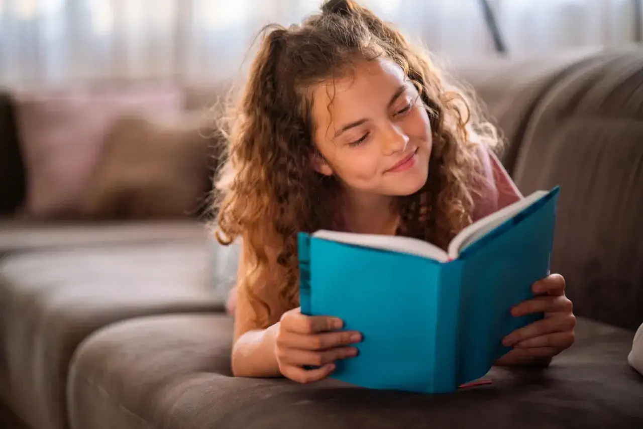 Ein M&auml;dchen liegt auf einem Sofa und liest ein blaues Buch. Sie strahlt und zeigt, wie sie eigenverantwortliches Lernen genie&szlig;t.