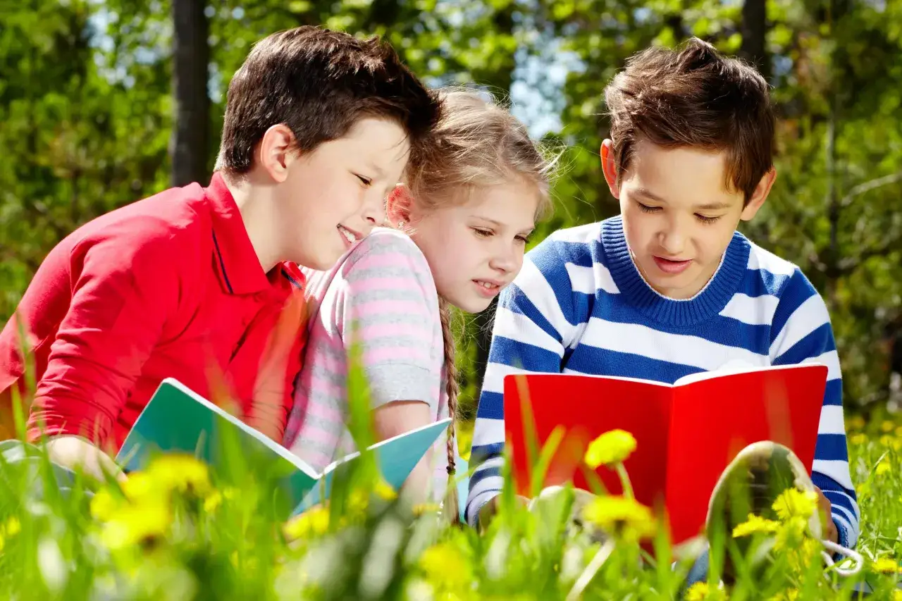Drei Kinder lernen im Gr&uuml;nen f&uuml;r ihr Sprachcamp Englisch. Sie sitzen im Gras und lesen in B&uuml;chern.