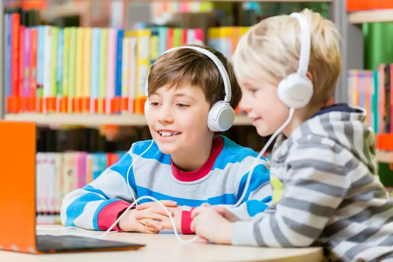 Zwei Jungen mit Kopfhörern lernen konzentriert am Laptop. Im Hintergrund bunte Bücherregale. Musik beim Lernen macht Spaß!