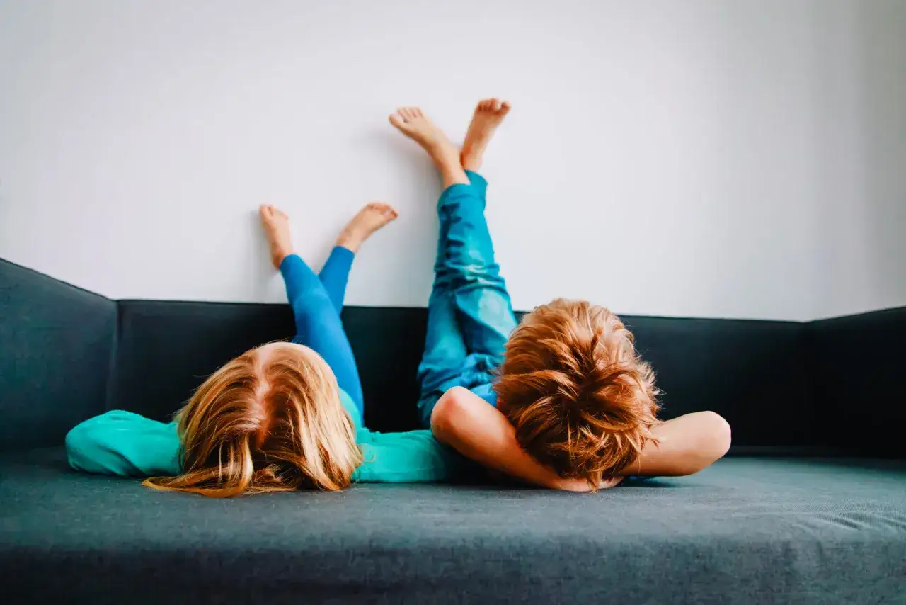 Zwei Kinder liegen kopfüber auf einem Sofa, die Beine an der Wand. Sie machen keine Hausaufgaben, sondern entspannen sich.