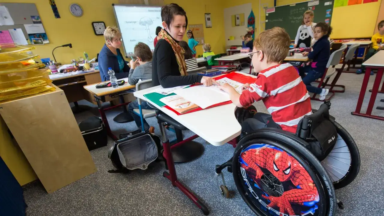 Lehrerin unterst&uuml;tzt Jungen im Rollstuhl beim Lernen. Ein Beispiel f&uuml;r gelebte Inklusion an Schulen, wo alle Kinder gemeinsam lernen.