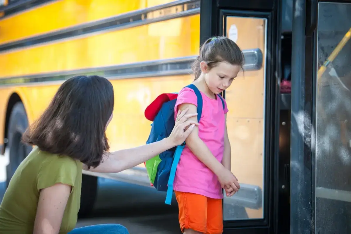 Ein kleines M&auml;dchen mit Rucksack steht am Schulbus. Ihre Mutter tr&ouml;stet sie, da das Kind Angst vor anderen Kindern hat.