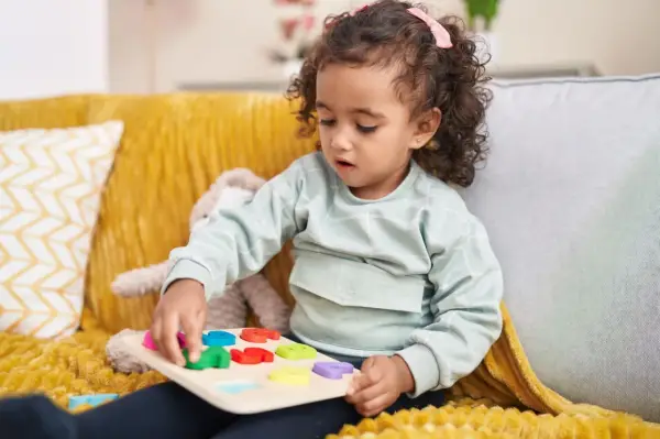 Kleines Mädchen mit Locken spielt konzentriert mit einem bunten Steckpuzzle. Das Spielzeug hilft, die **Konzentration zu steigern**.