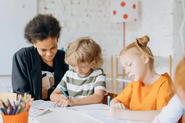 Kinder konzentriert bei Lernritualen in der Grundschule. Ein Mädchen mit schwarzer Afro-Frisur schaut zu, während ein Junge mit blonden Locken und ein Mädchen mit roten Haaren schreiben.