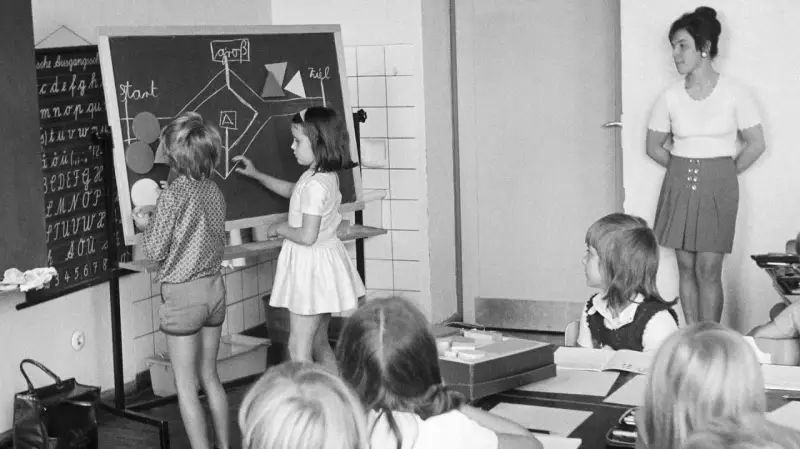 Unterricht in einer Schule um 1900. Kinder lernen an einer Tafel, während die Lehrerin zusieht.